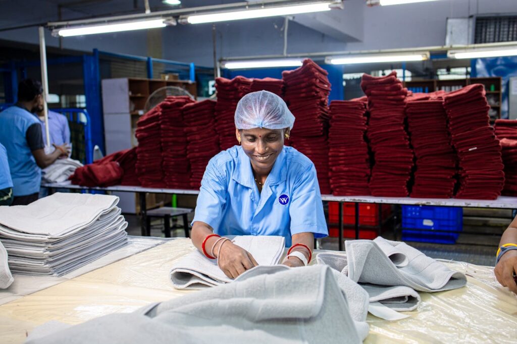 A woman working in a textile factory, folding fabric with stacks of cloth in background.